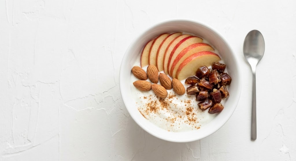 Greek yogurt topped with sliced apples, almonds, and dates on a white breakfast plate.
